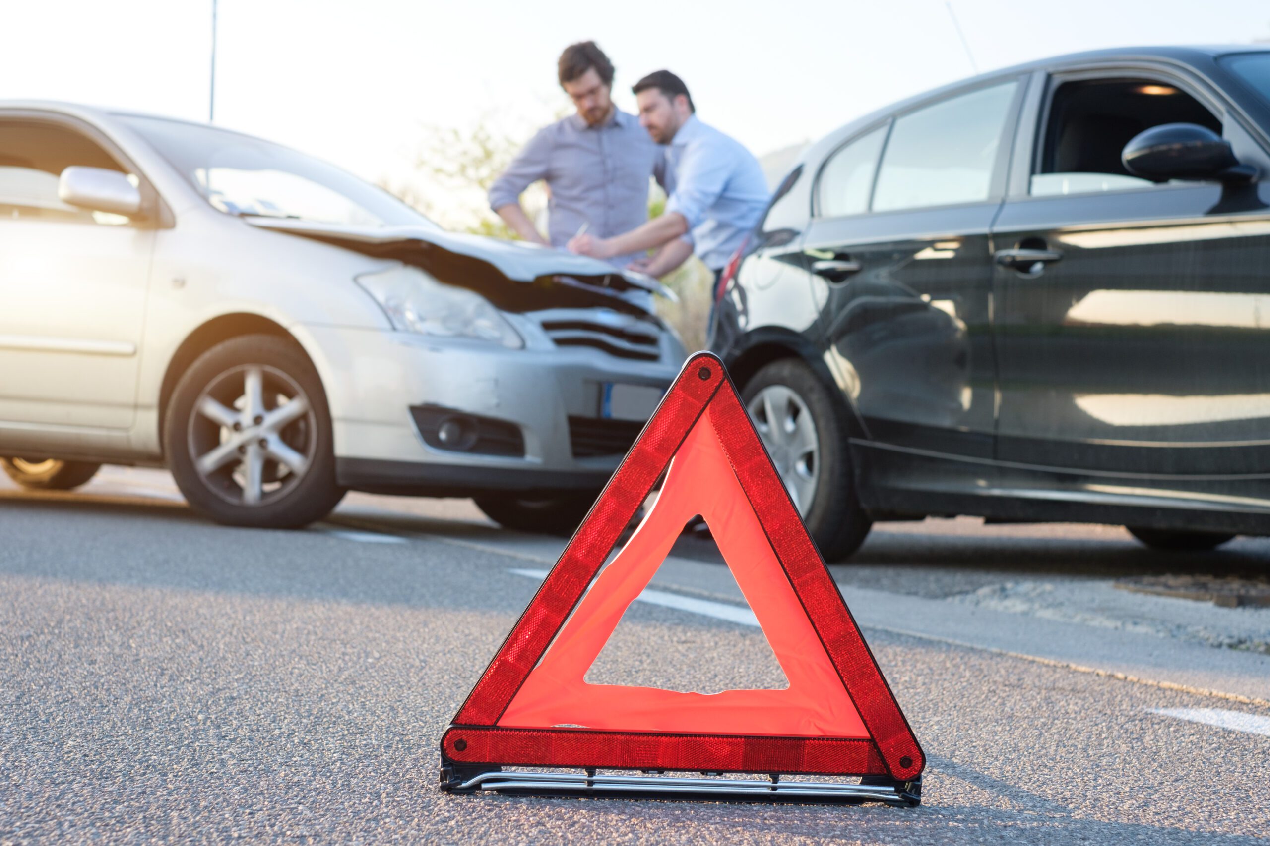 Two men in professional attire stand in the background of a car accident, discussing damage to a white vehicle.