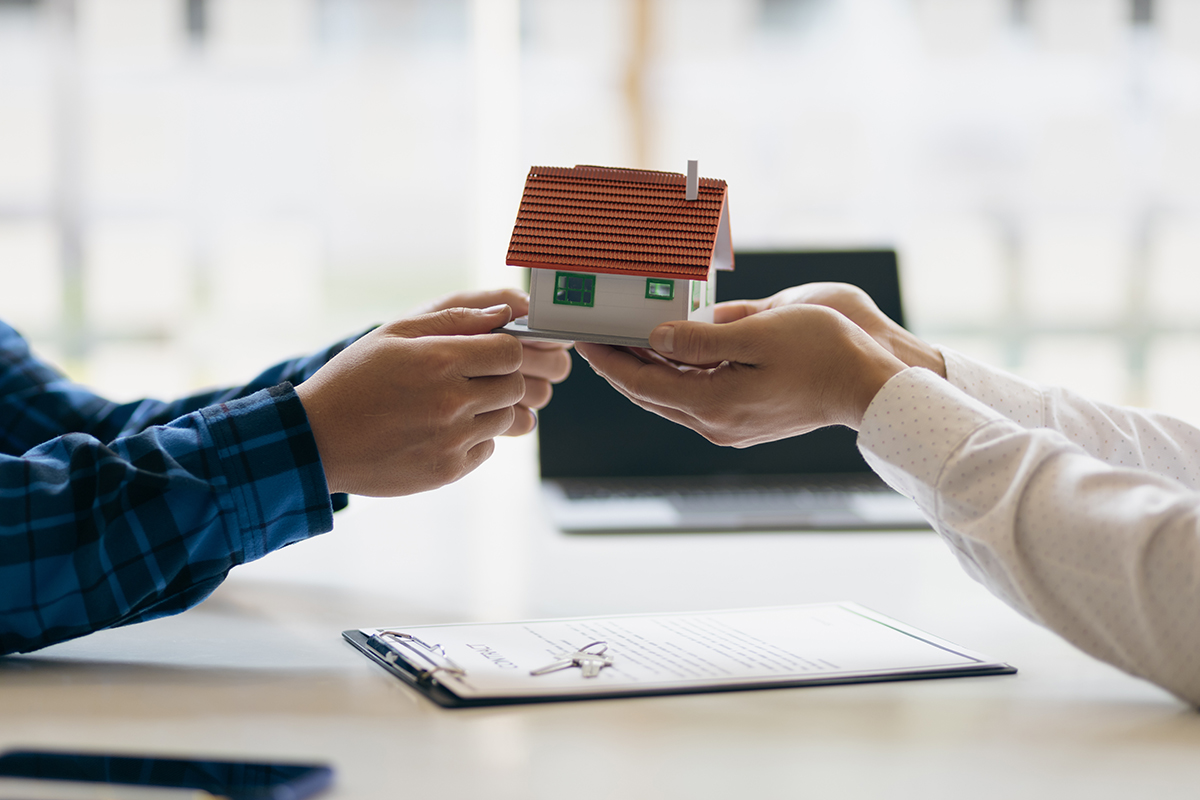 Two people are holding a small model of a home, and it looks like one person is receiving the model from the other.