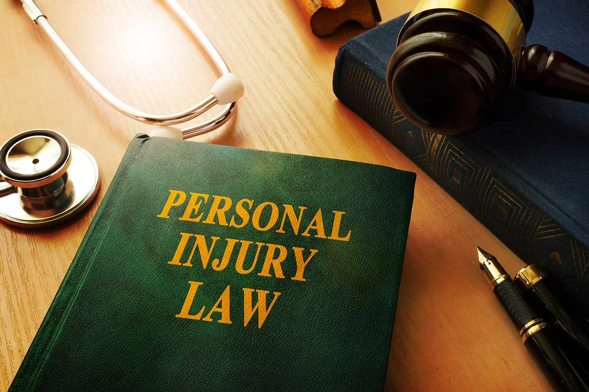 A green book with yellow writing on it that says “personal injury law” is surrounded by a stethoscope, a pen, and a blue book with a gavel resting on it.